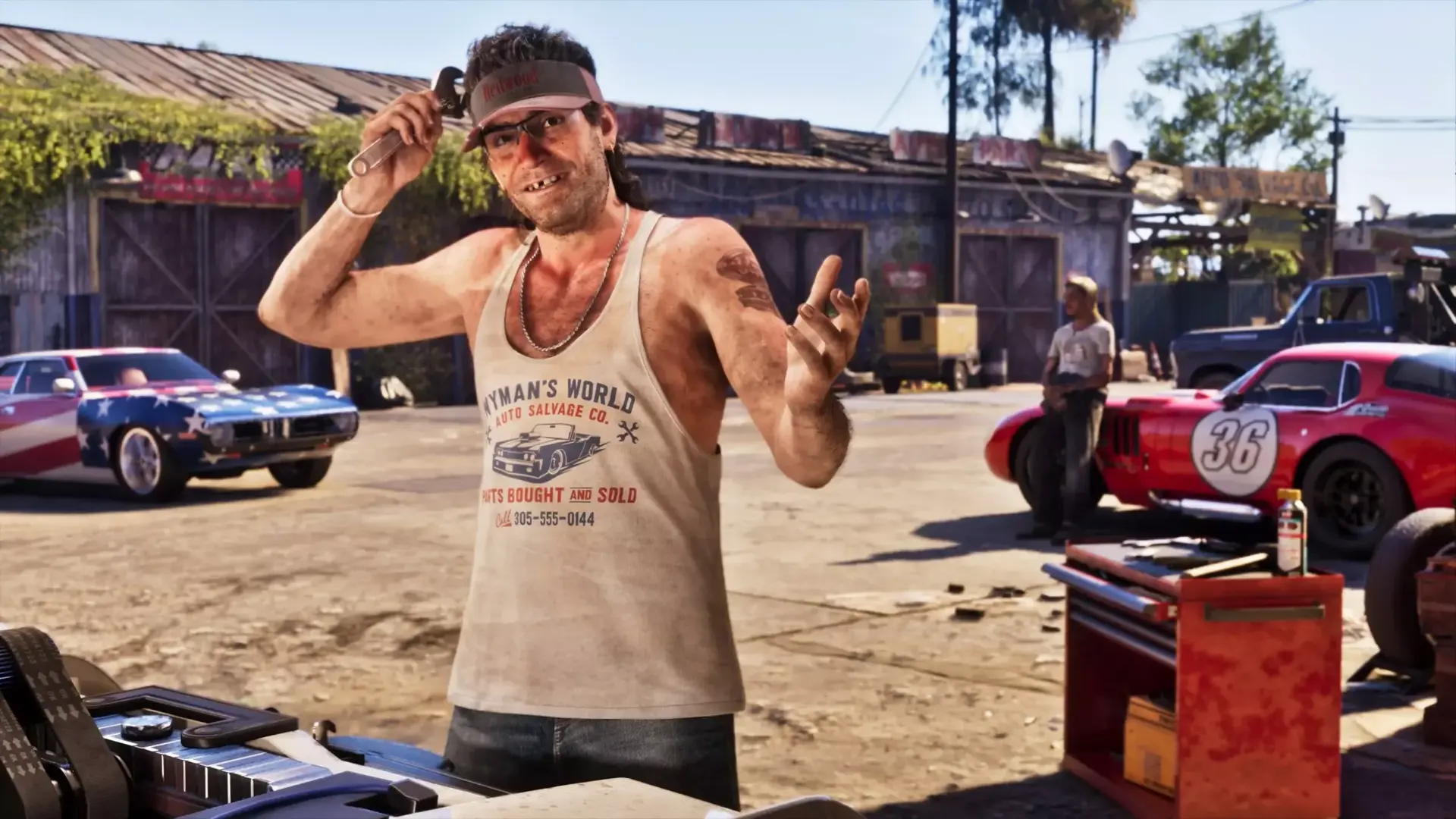 A man in a visor and tank top smiling in a yard of classic project cars
