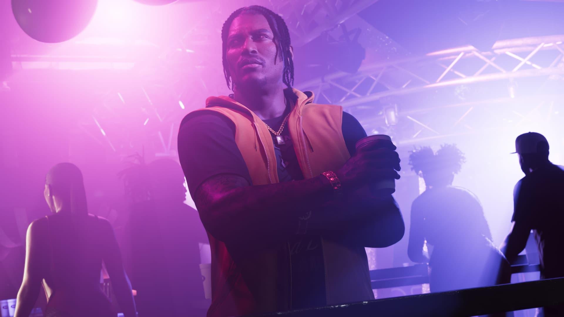 Dre'Quan Priest con un gilet arancione con cappuccio e le braccia conserte in un locale illuminato di viola, in uno screenshot ufficiale di GTA 6