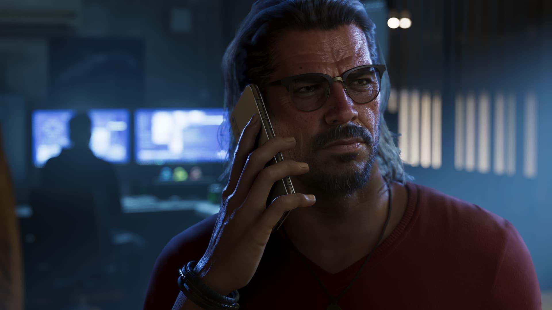 Raul Bautista mit Brille und langen Haaren hält sich in einem dunklen Raum ein Handy ans Ohr, hinter ihm Monitore, in einem offiziellen GTA-6-Screenshot